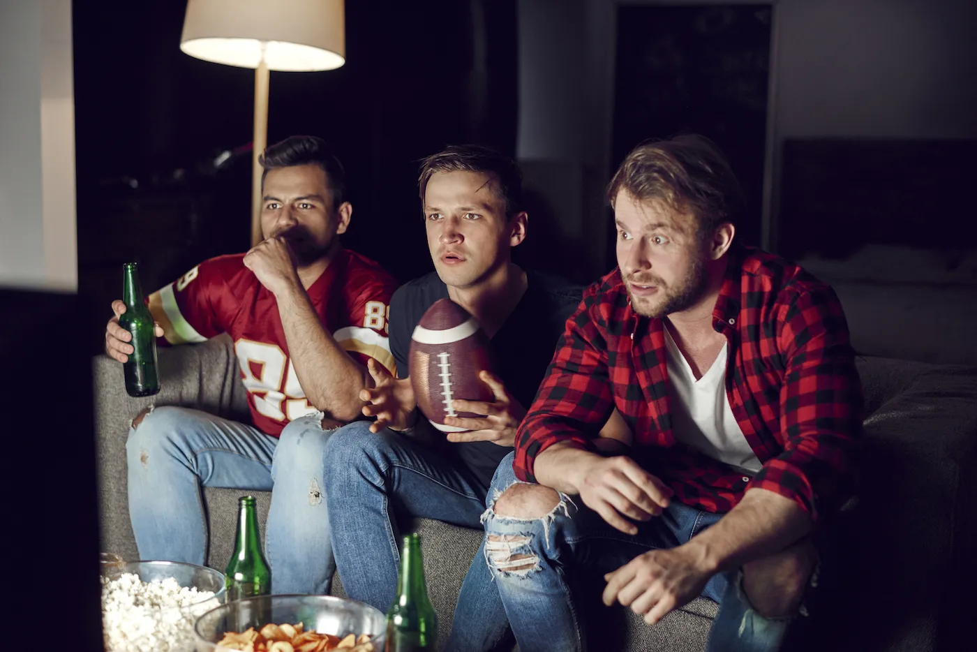 Three football fans sitting on the couch watching the game with beverages and snacks.