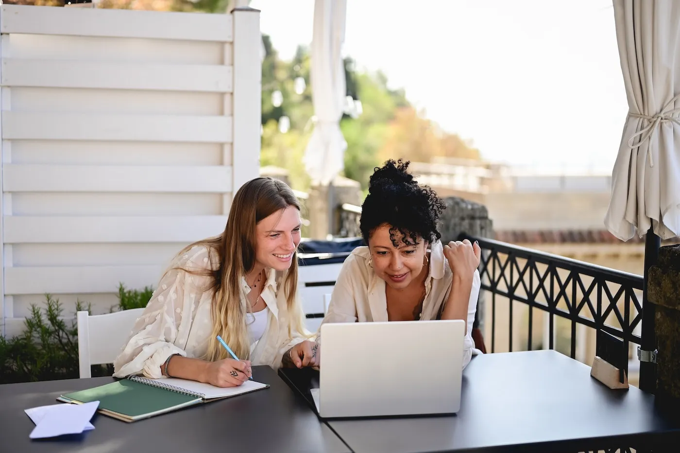 Two cheerful women using laptop on a beautiful outdoor balcony.