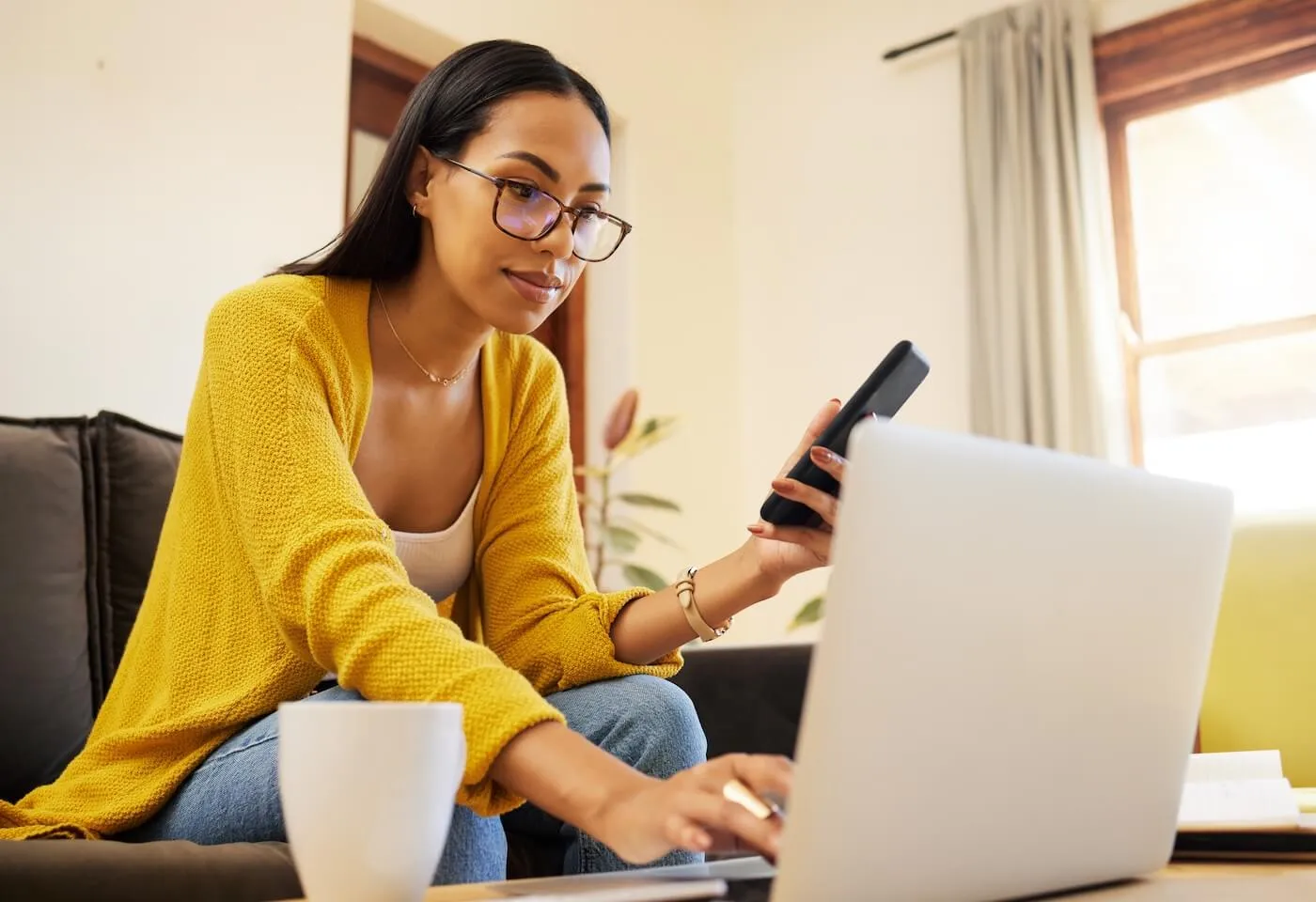 Focused young woman using her laptop while holding a smartphone