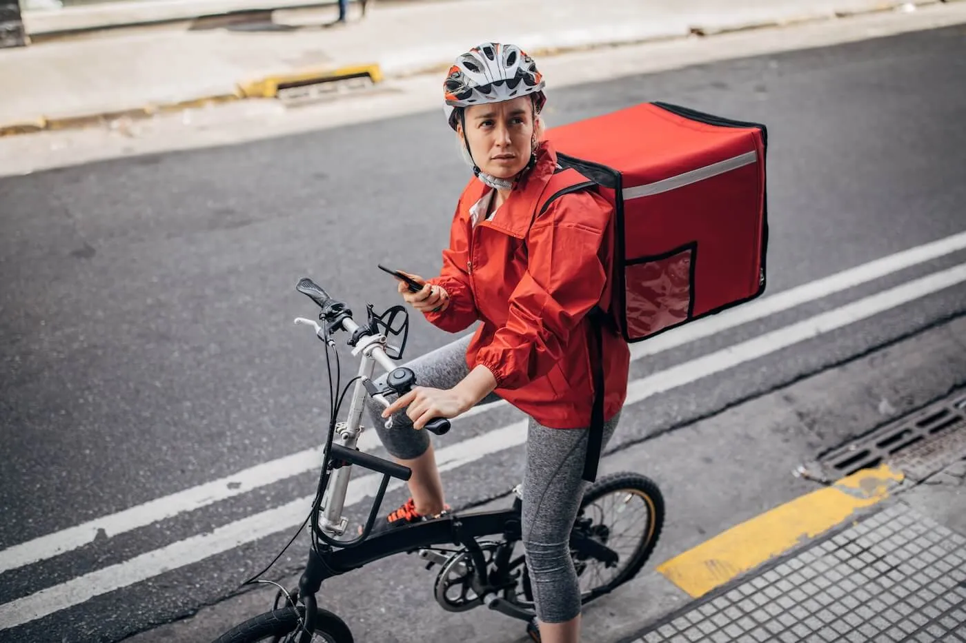 A delivery woman on the bicycle with the food delivery backpack is checking the address on her smartphone