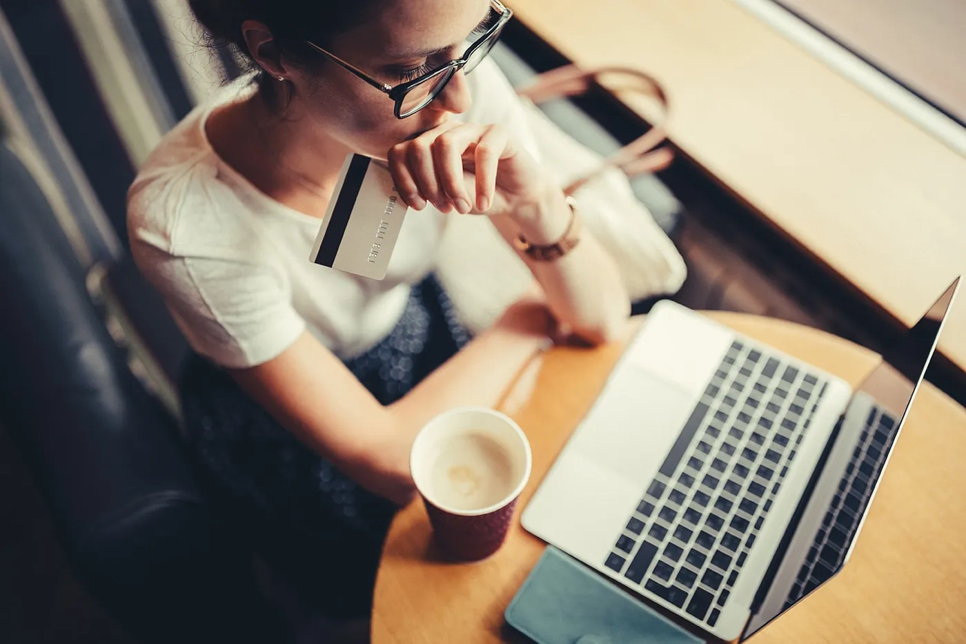 A woman sitting in a coffee shop holding her credit card, going over credit card payments on her laptop.