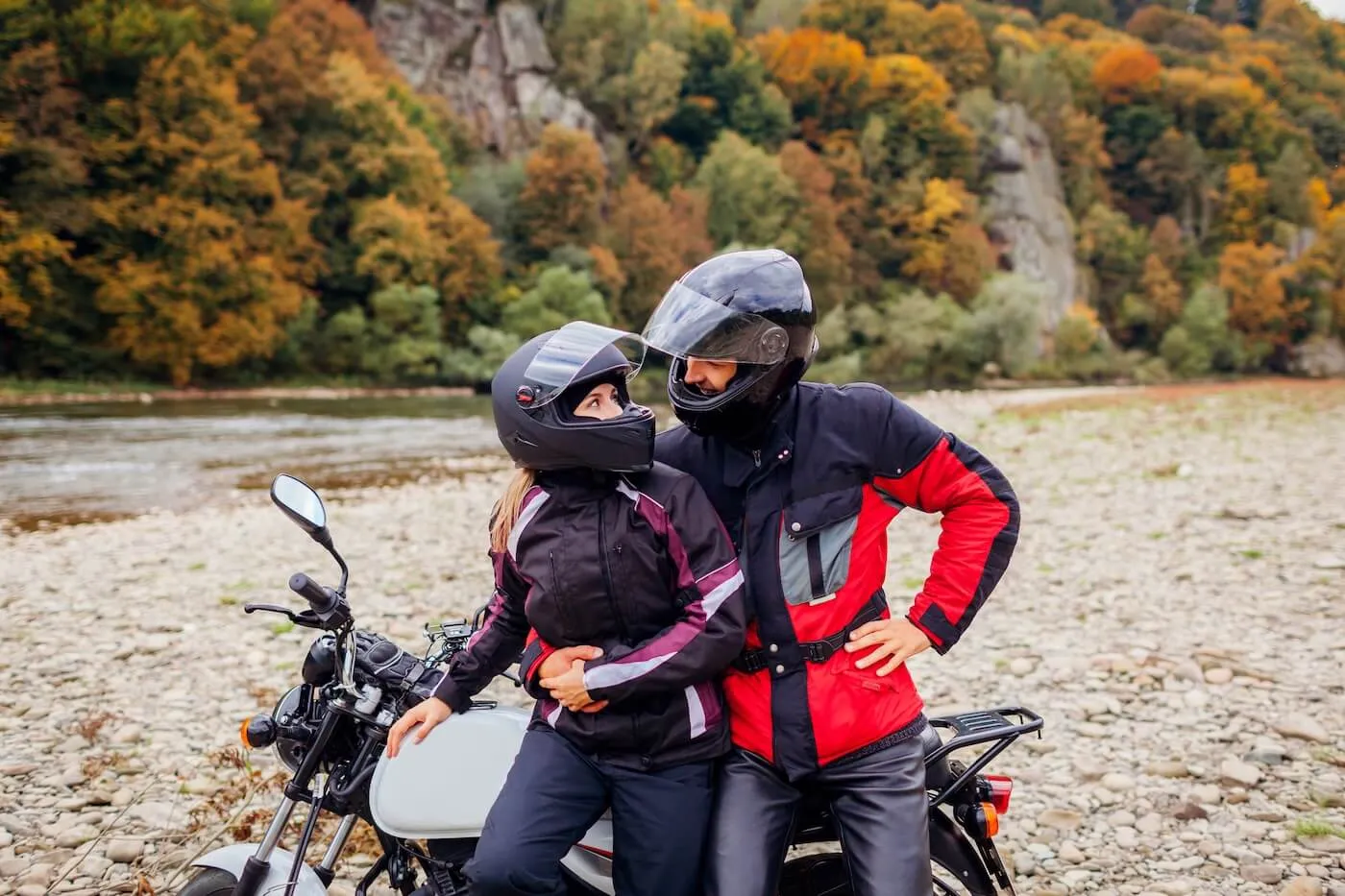 Happy couple in helmets hugging each other next to their motorcycle, a mountain river on the background