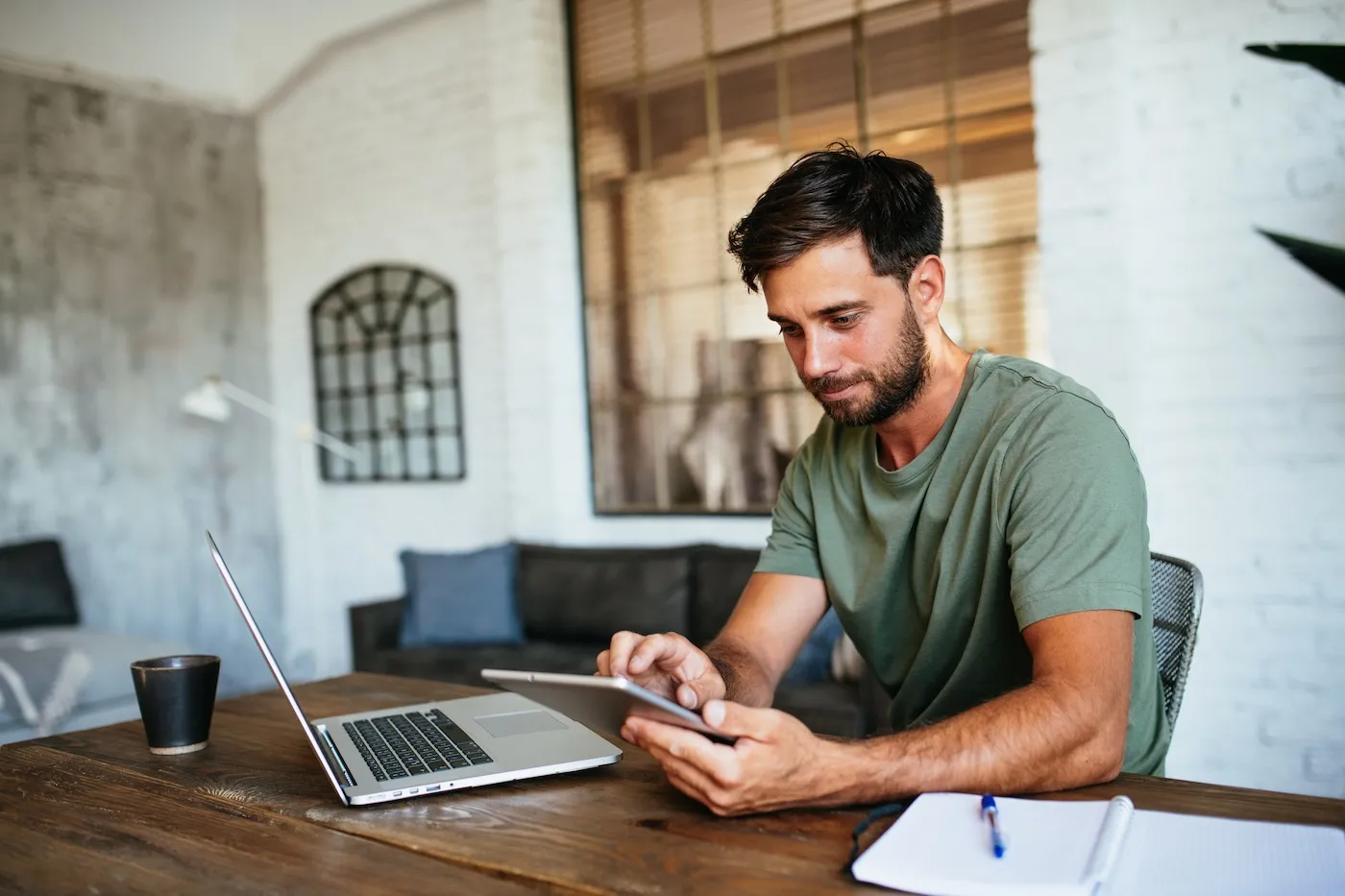 Young man working from home, focused on apps and software on laptop and digital tablet.