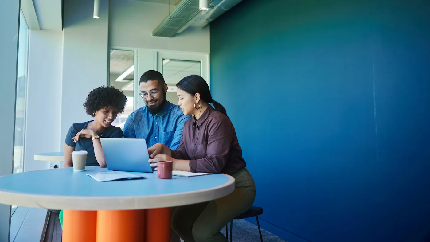 A group of a smiling young businesspeople working together on a laptop during a meeting around a table in an office lounge