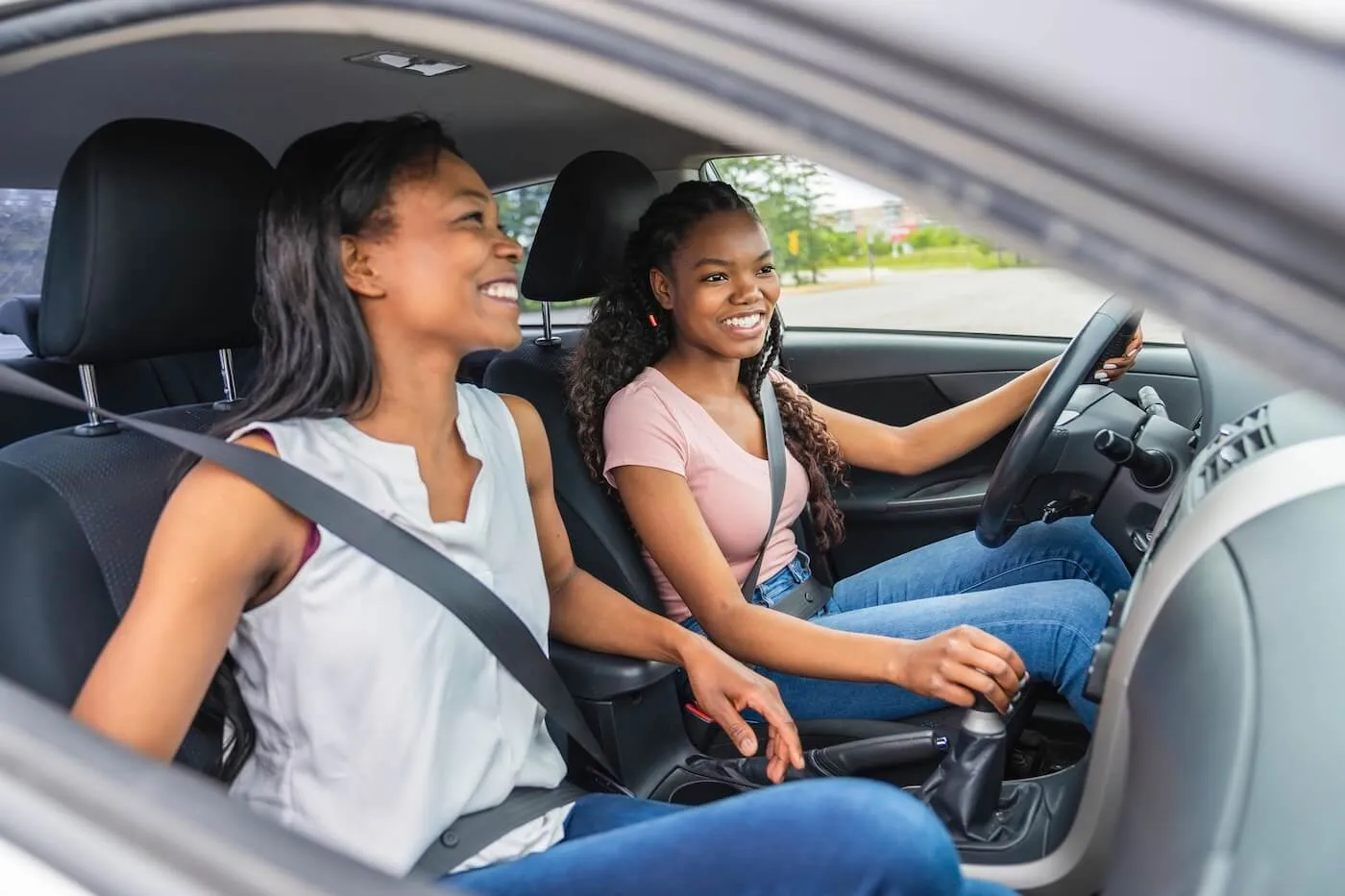 A smiling teenage girl is driving a car while her mother is sitting on a passenger seat