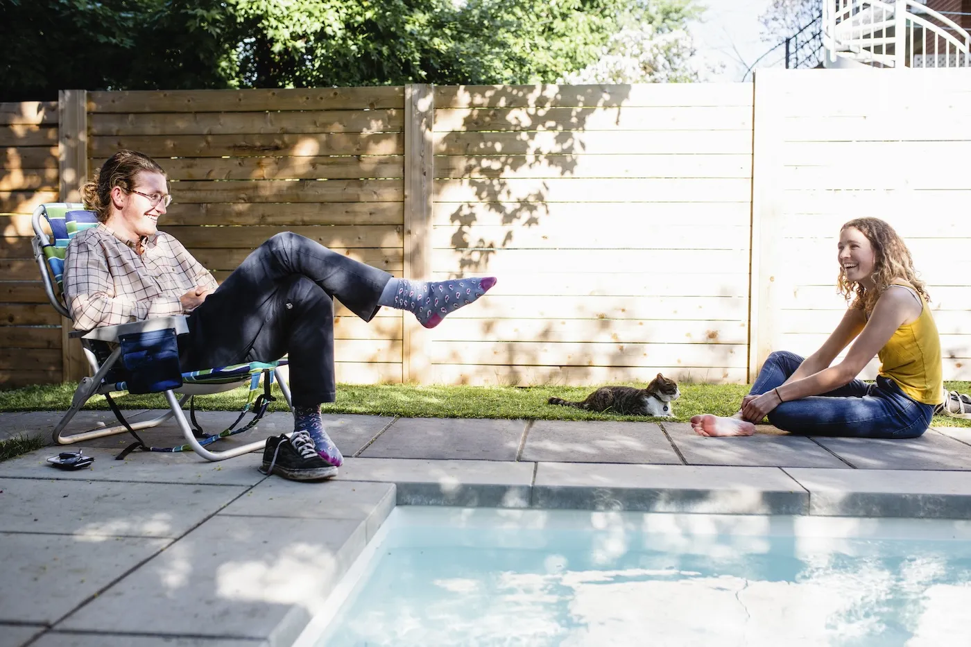 A man and a woman relax by their backyard pool with their cat.