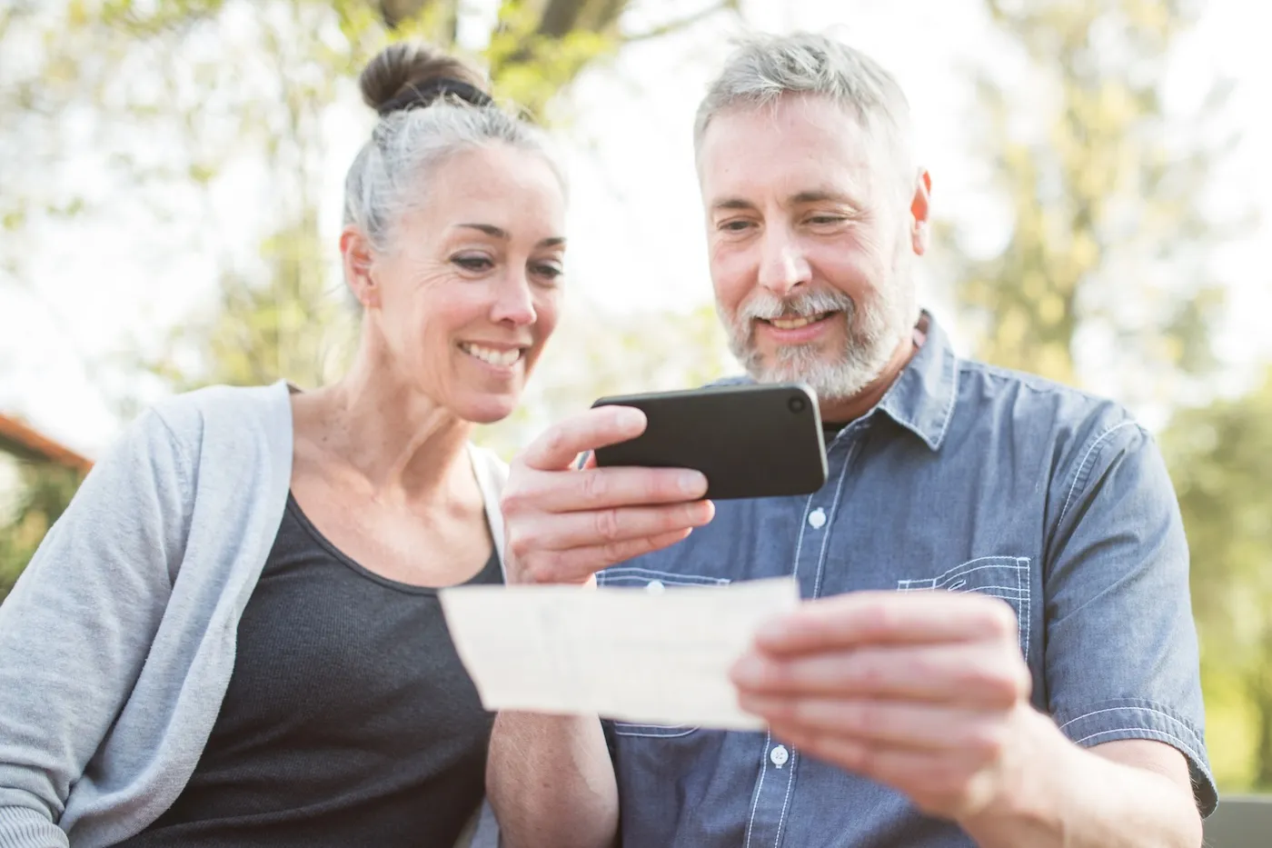 A couple enjoy time with each other in a beautiful outdoor setting, the sun casting a golden glow on the scene. The man takes a picture of a check with his smart phone while checking his bank account.