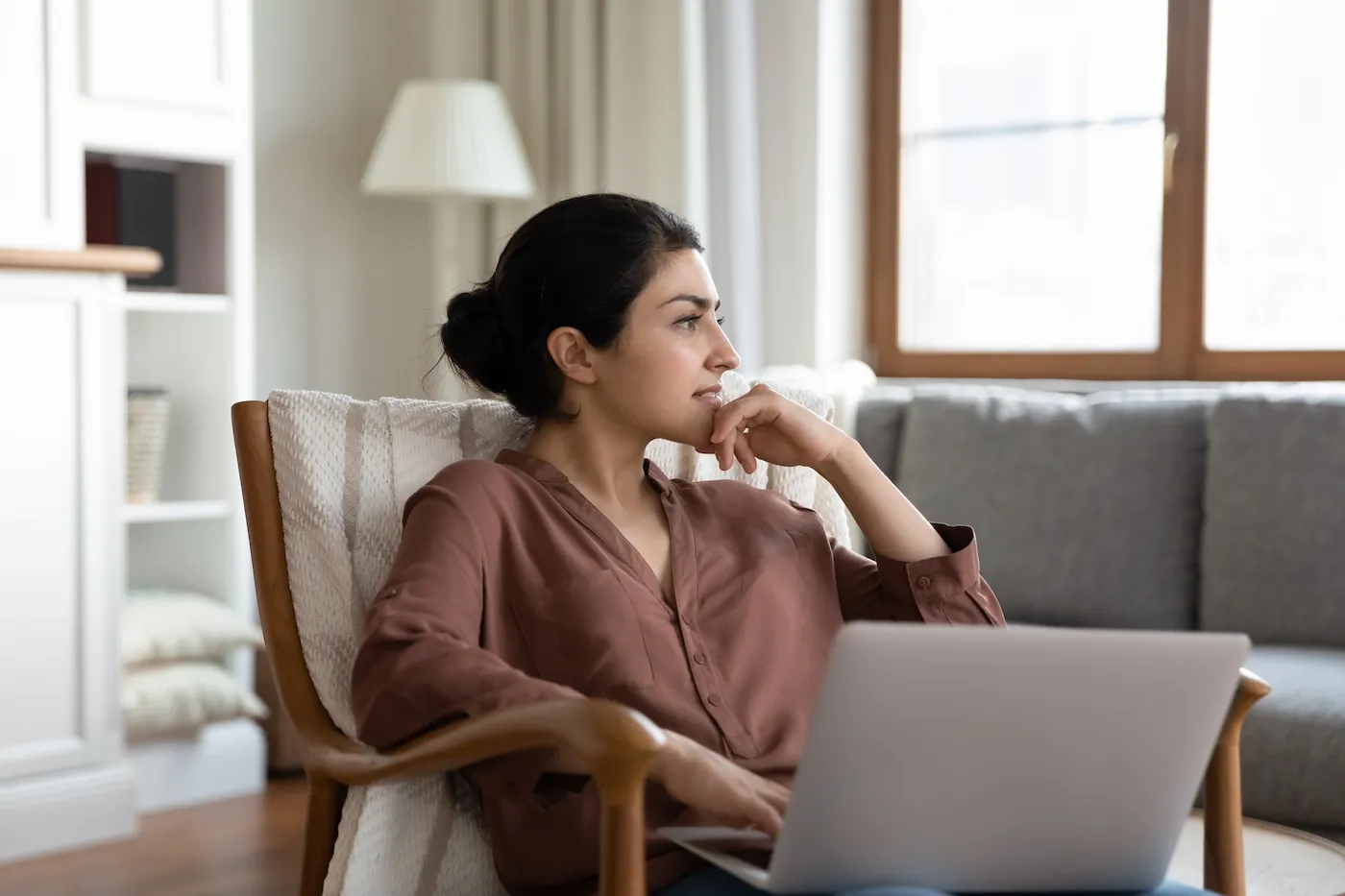 A thoughtful young woman resting on a cozy seat in her living room with her laptop, deciding whether to make a purchase.