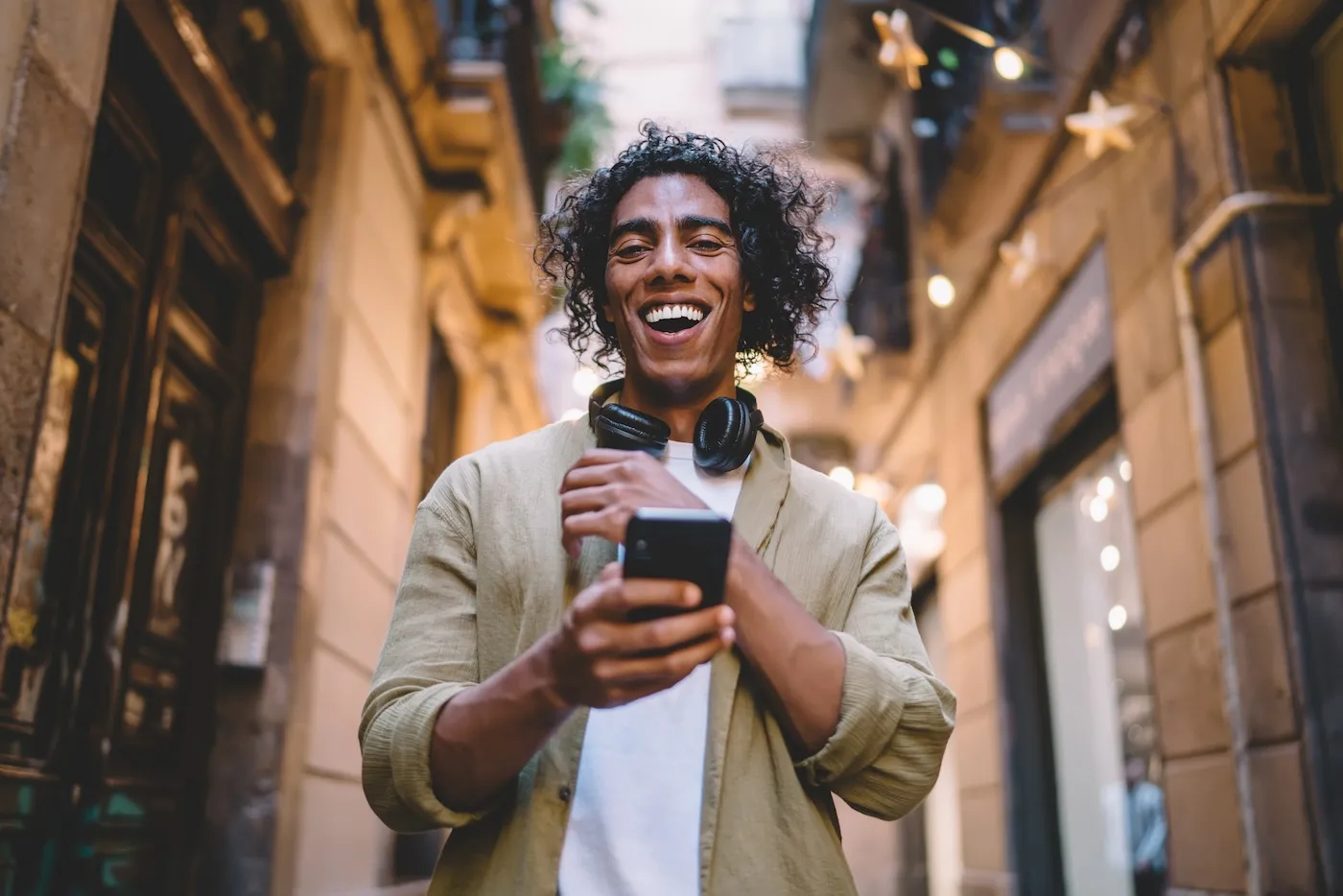 A smiling man in casual clothes with wireless headphones browsing smartphone while standing on narrow street between stone buildings in daytime.