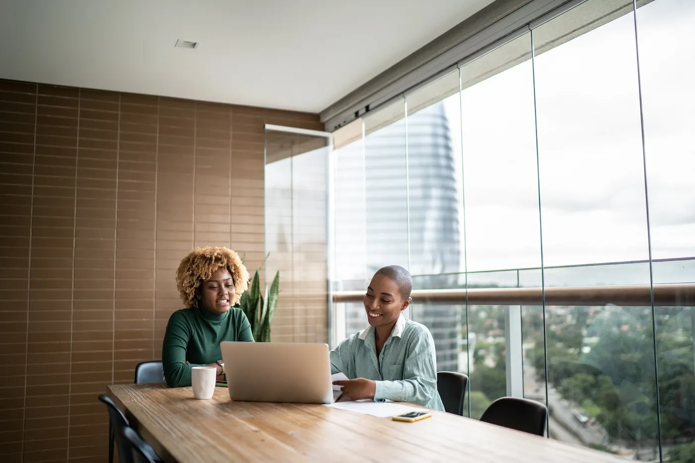 Two women sitting at a table in a room with large windows, using a laptop and discussing finances.