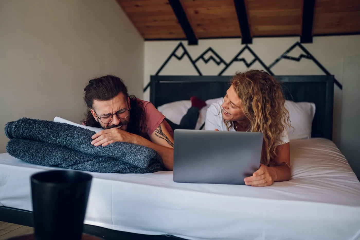 Young couple lying in a cozy bed and having a conversation while using a laptop.