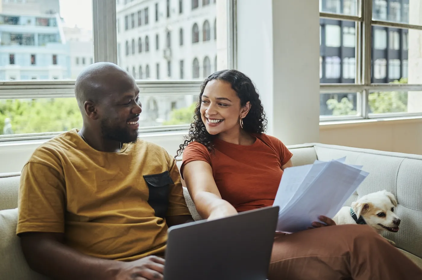 A young happy couple working on their budget and finances on a sofa from home with their dog using a laptop computer.