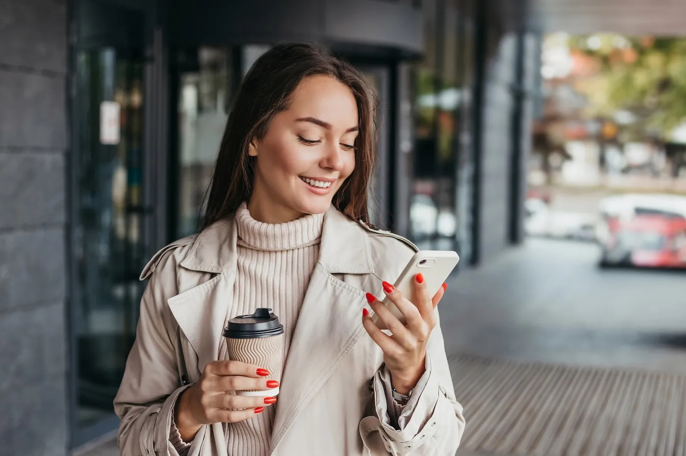 Young smiling woman holding a smartphone and a cup of coffee looking at device screen against the background of an office building.