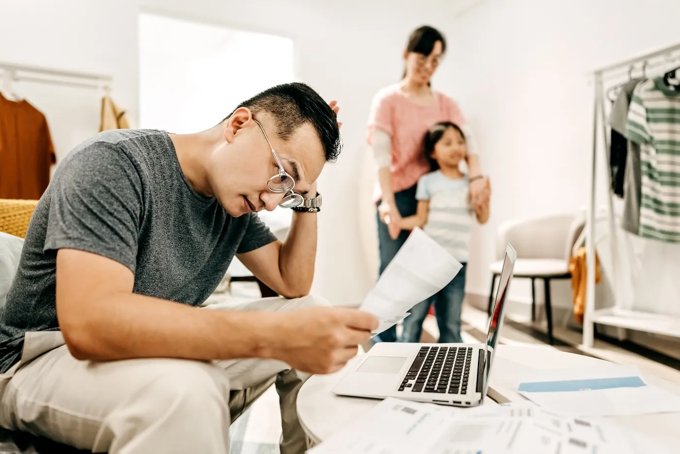 Man with eyeglasses working on finances at home in the living room while a woman and her daughter are playing.