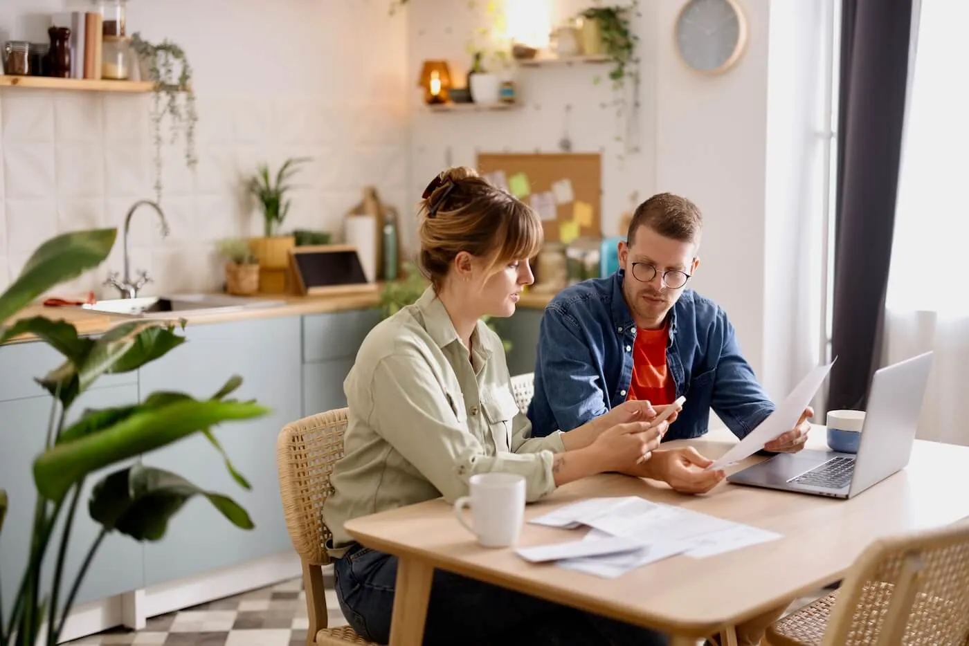 A young couple compares home insurance plans using laptop and printouts while having hot drinks in their kitchen