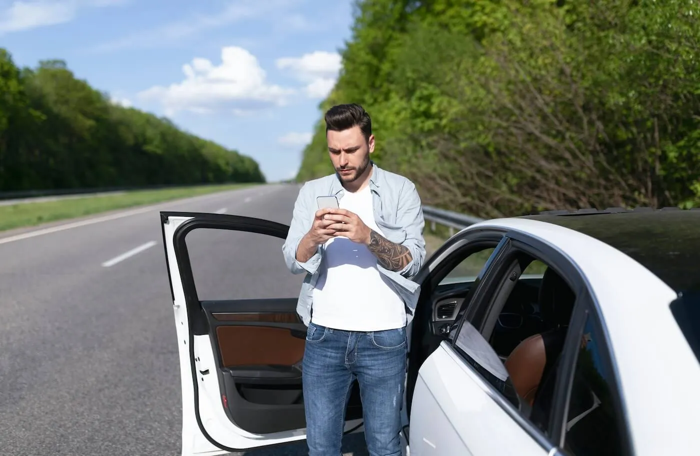 Concerned man making using his smartphone while standing next to an open car on an empty countryside road