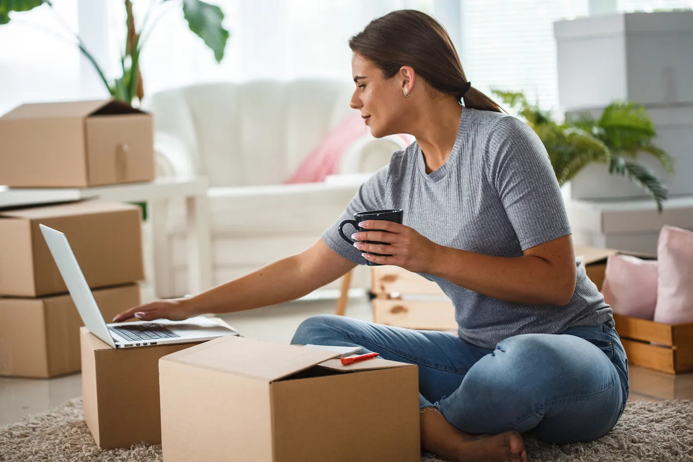 Side view of a young woman sitting on floor of the living room, surrounded by boxes. She is drinking coffee and using laptop.