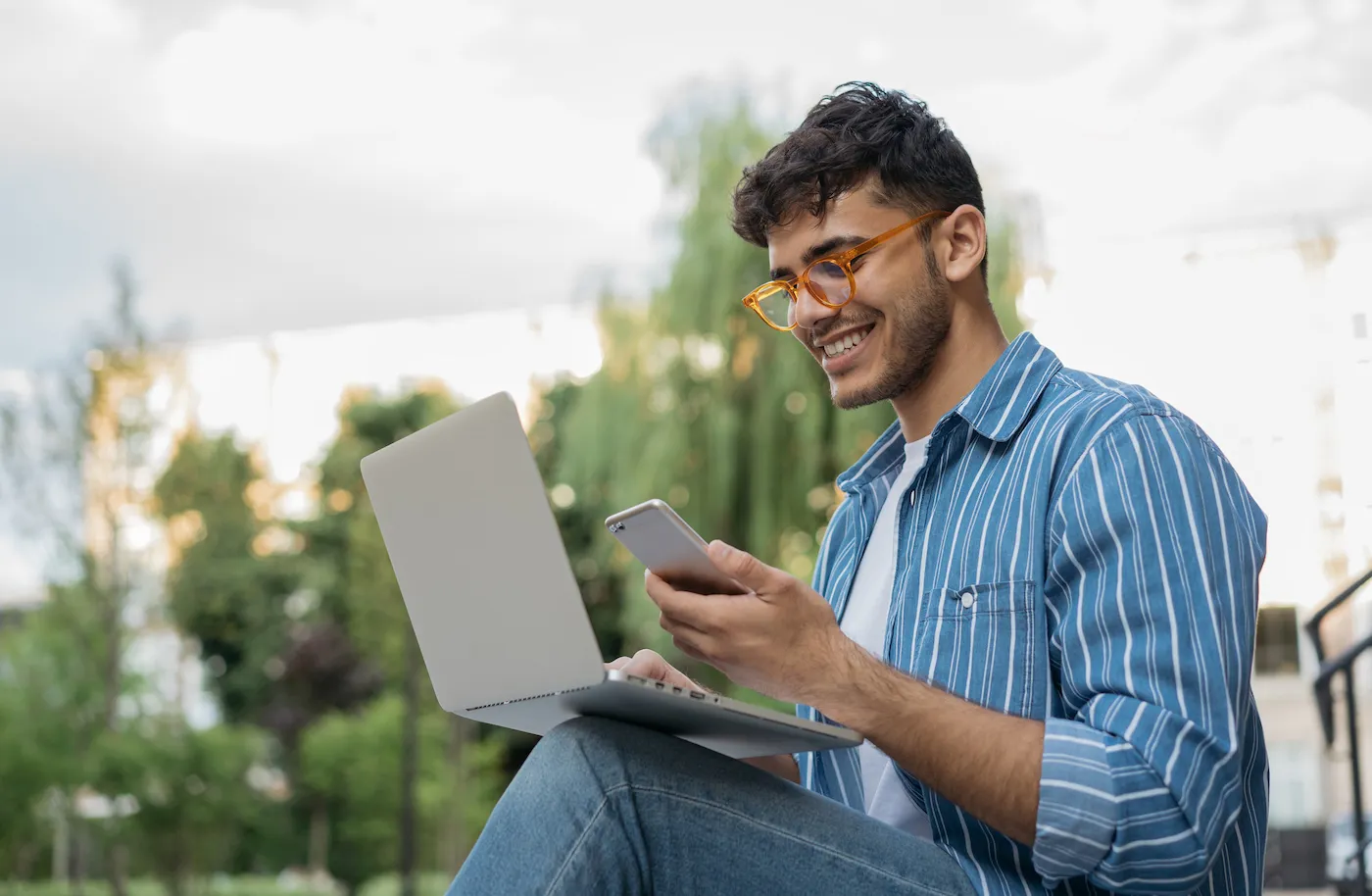 A man getting an overdraft fee refund, sitting outside using his laptop and mobile phone.