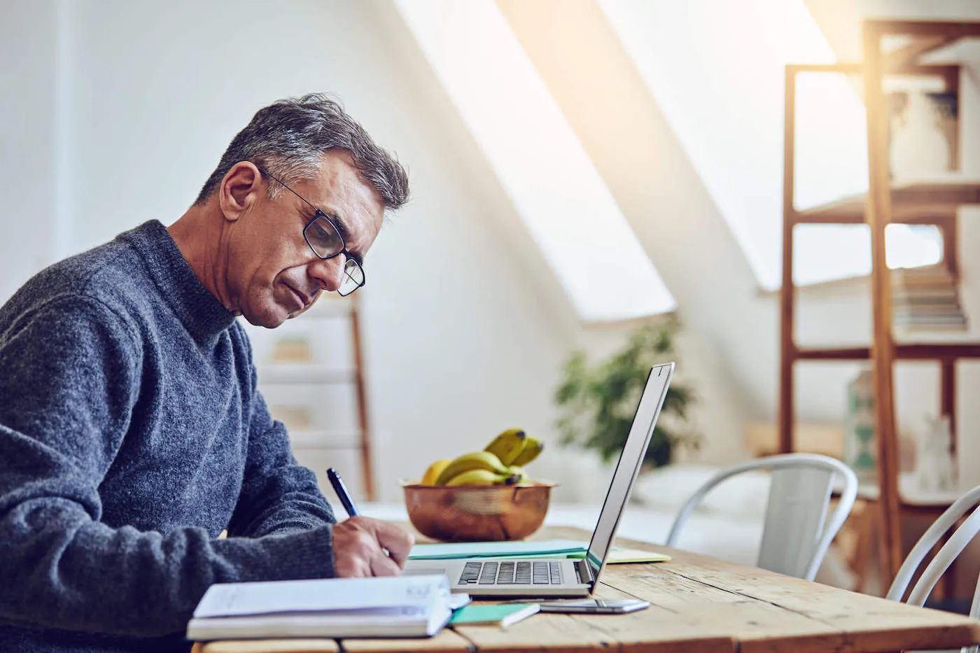 Man in glasses writing on paper while seated at a desk with an open laptop.