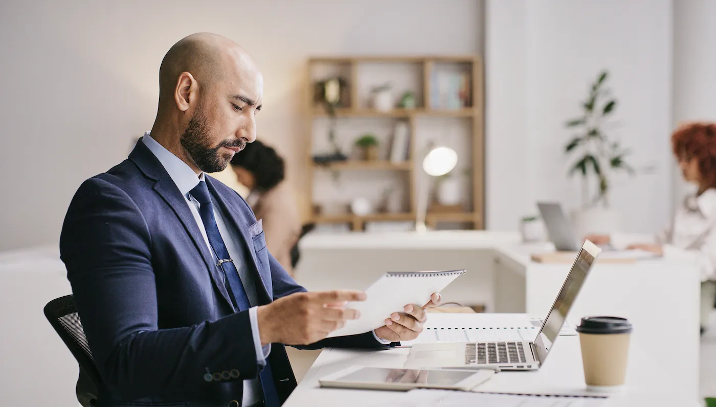 Man sitting at his desk looking at his savings account on a laptop, holding a notebook.