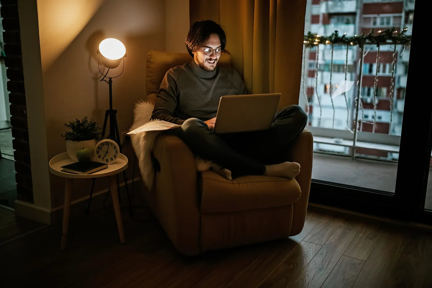 Man sitting cross legged in a comfy armchair in dark living room, looking at laptop and smiling