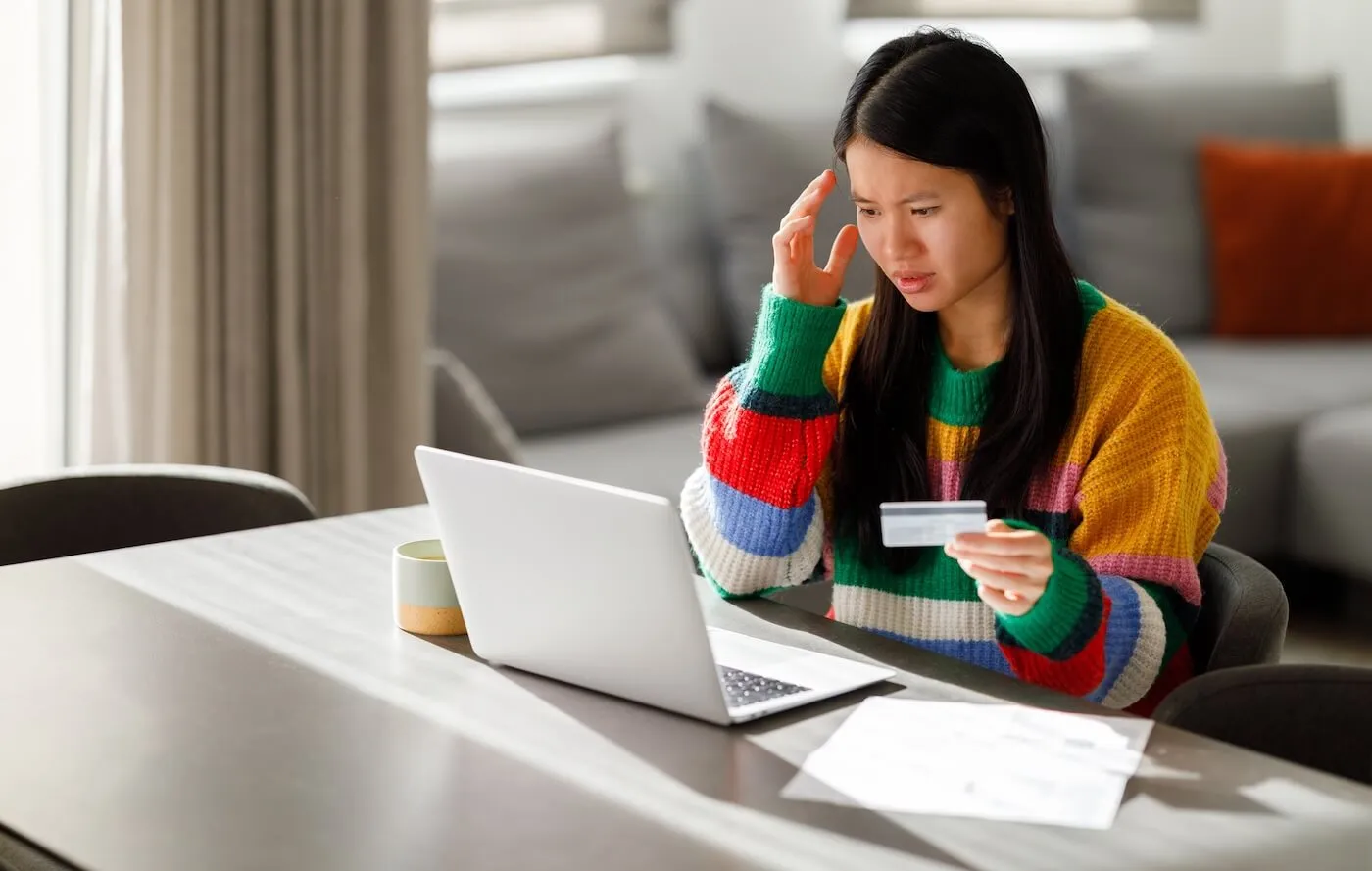 Concerned young woman is holding her credit card while looking at the laptop screen