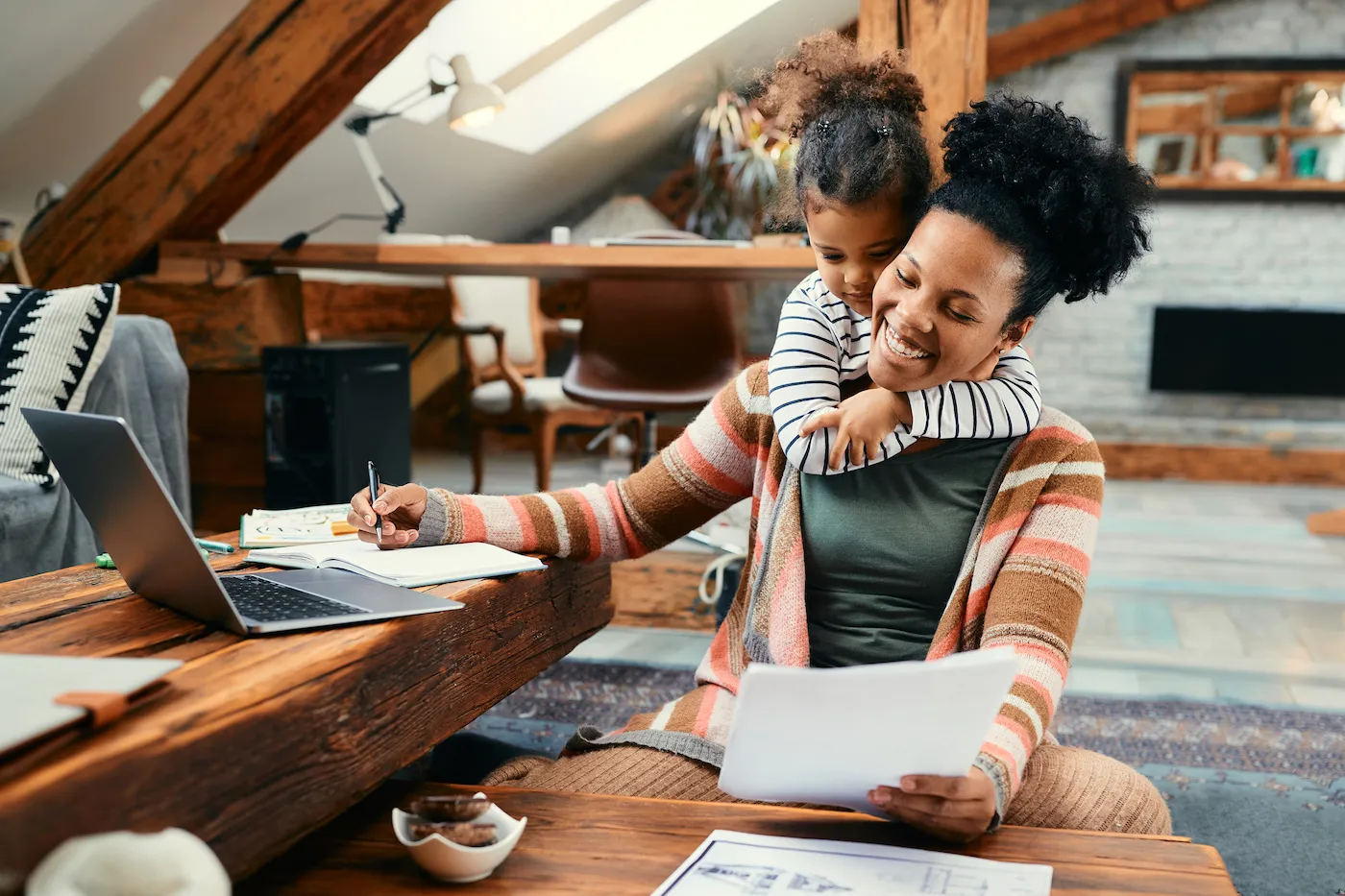 Happy mother using laptop and working on taxes while her daughter is embracing her at home.