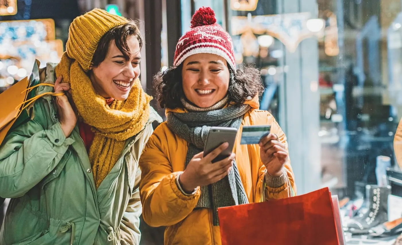 Two cheerful female friends in winter outfit holding shopping bags while making a payment through smartphone