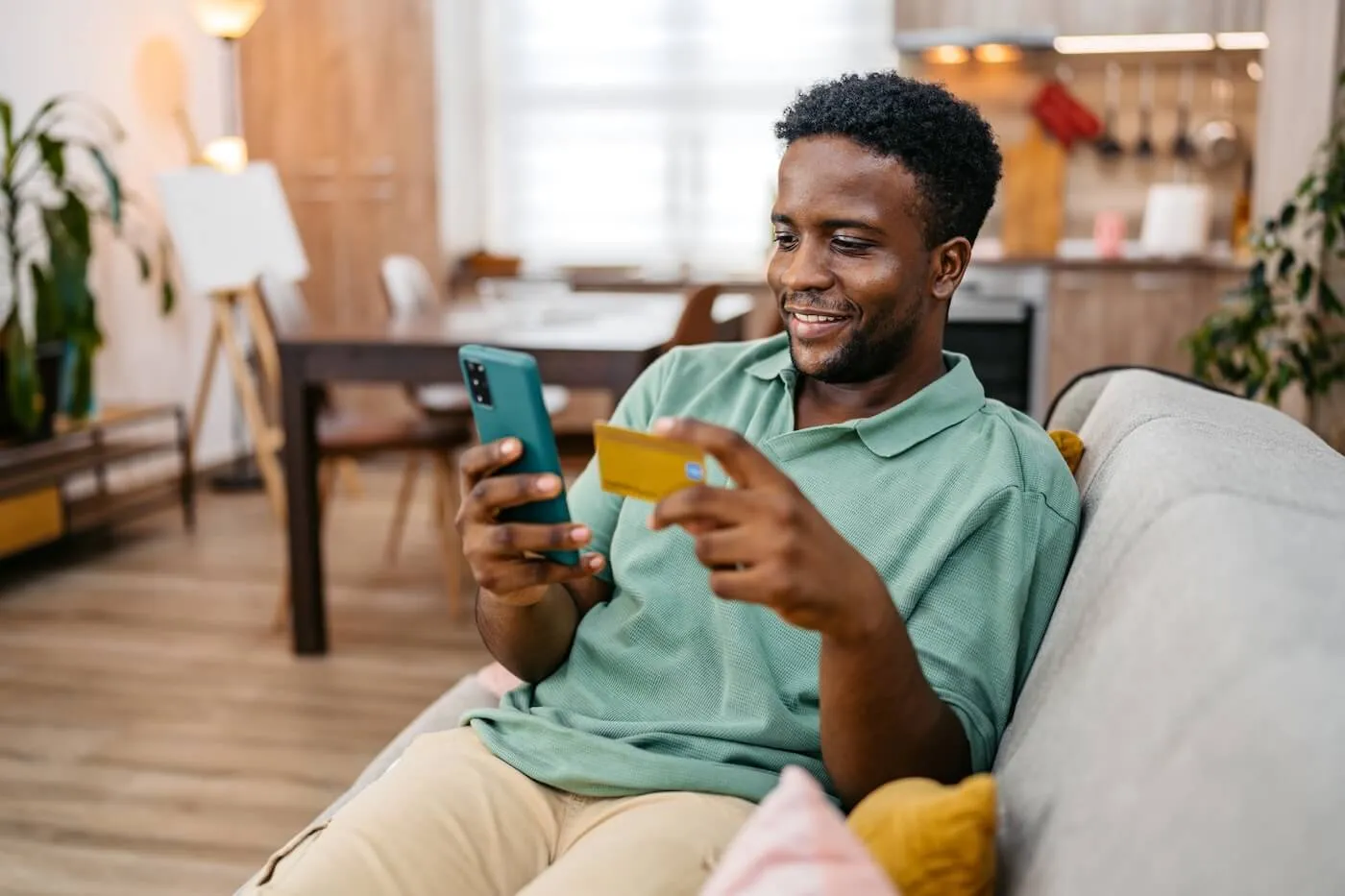 Smiling man is sitting on a couch and checking his credit card balance on his smartphone