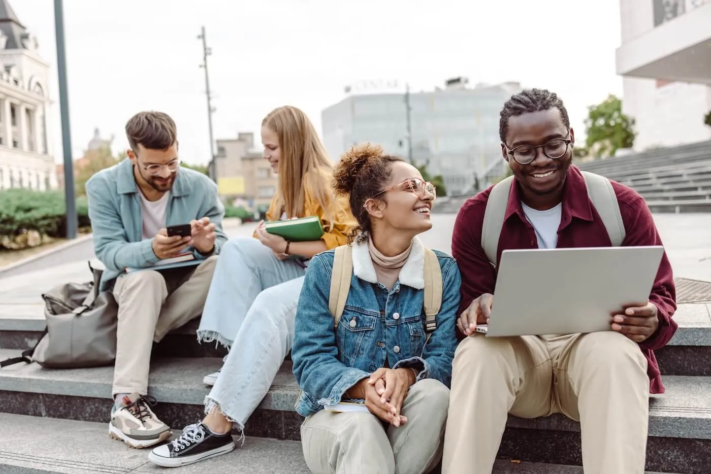 Four happy students, males and females, sitting on the stairs, chatting, and using their mobile devices