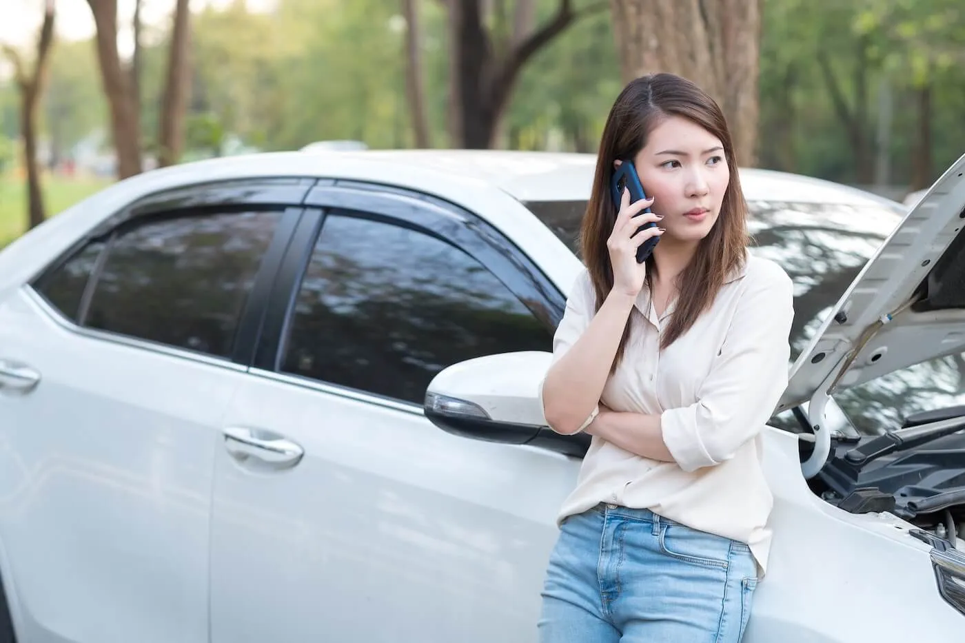 Concerned young woman making a phone call while standing next to a broken white car