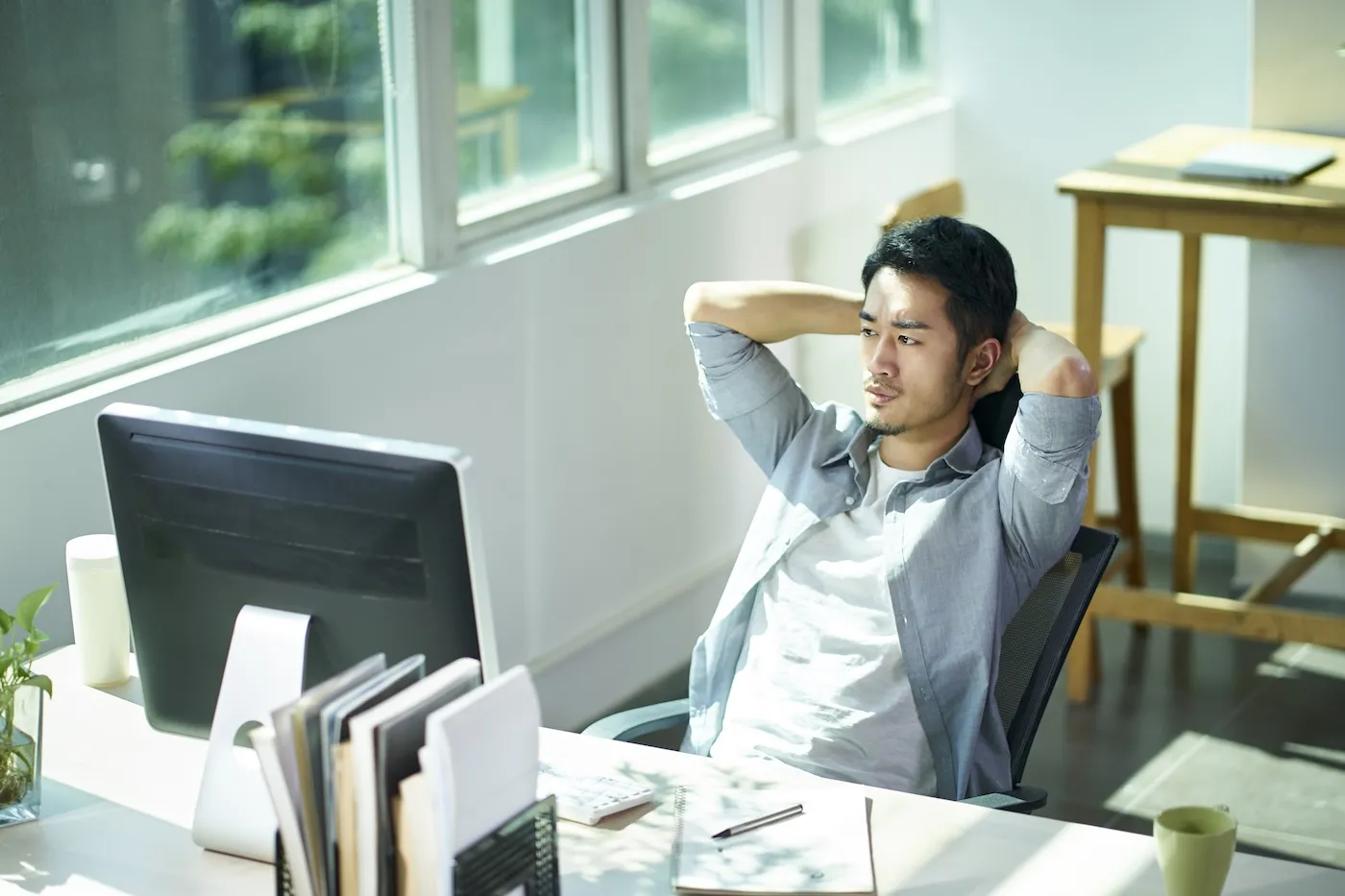 A worker contemplating in office, hands behind head frowning.