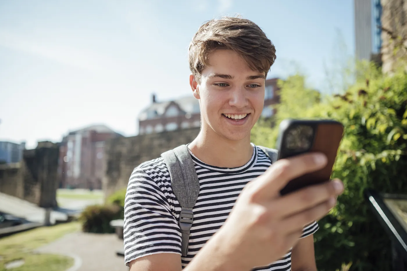 A university student using his phone and standing in a sunny public park in Newcastle Upon Tyne, North East England.