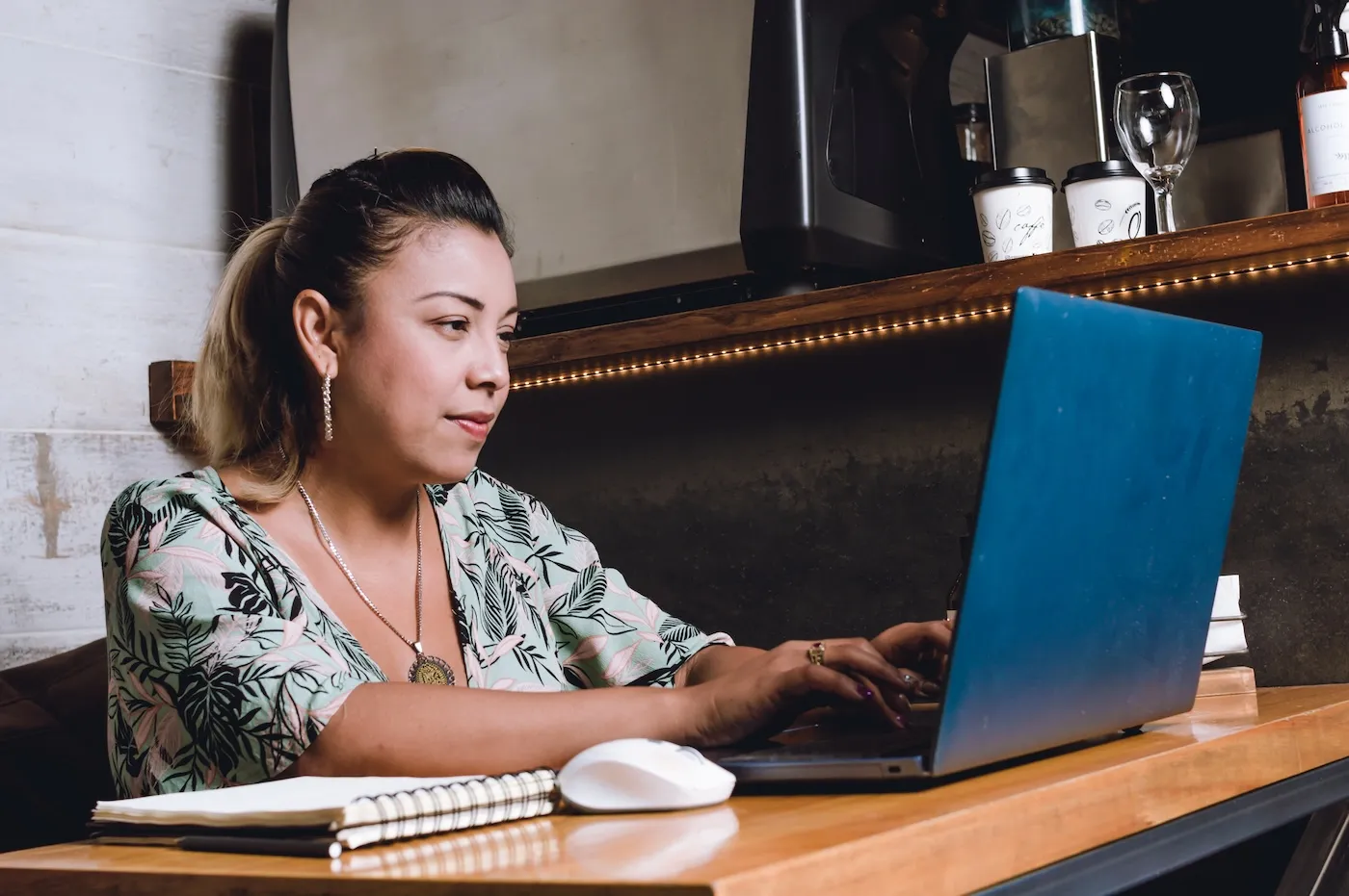 A woman working and typing on her computer using a coffee shop as her office with a notepad on the table.