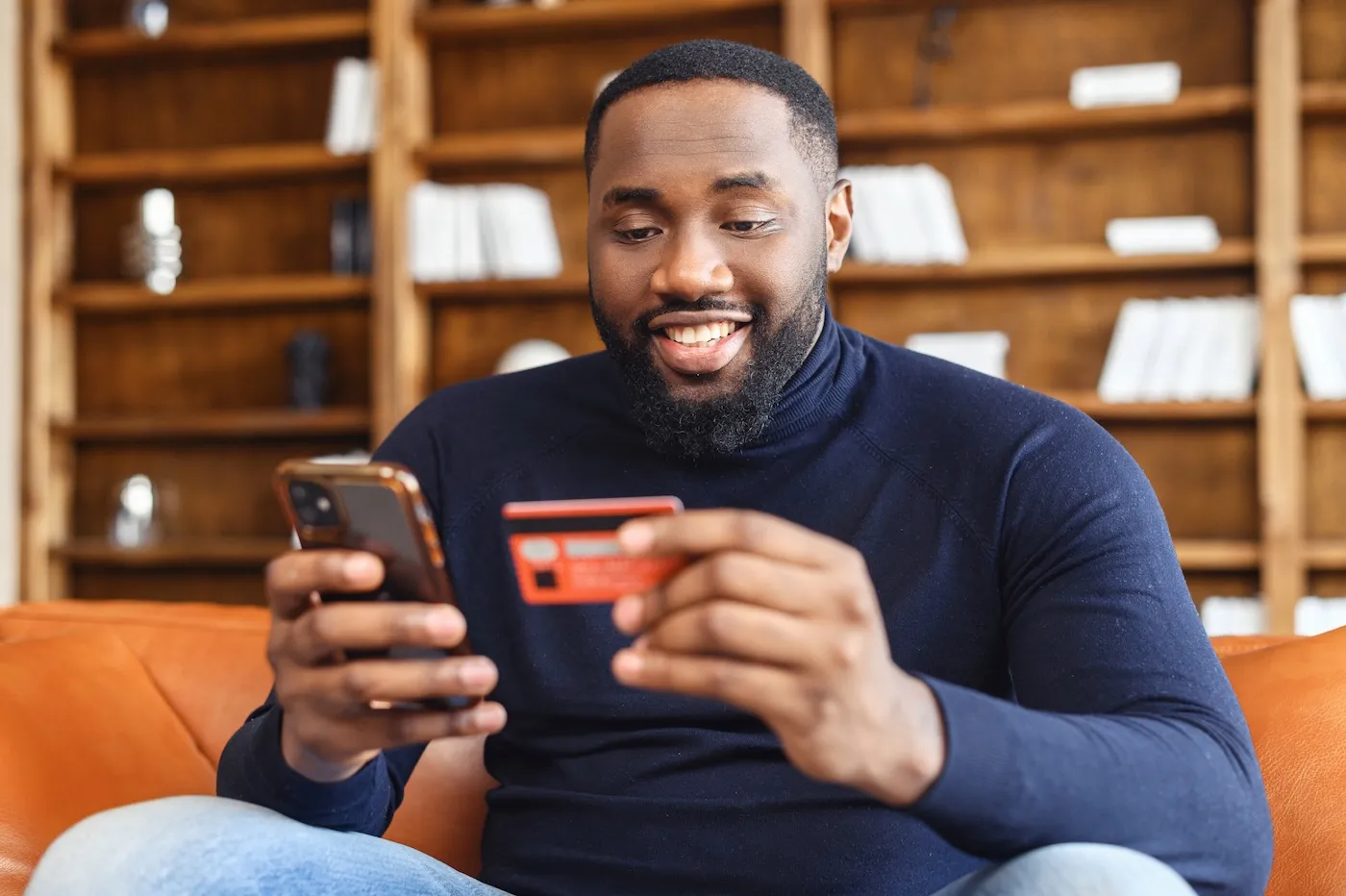 Smiling man shopping online sitting on the sofa at home, holding credit card and smartphone.