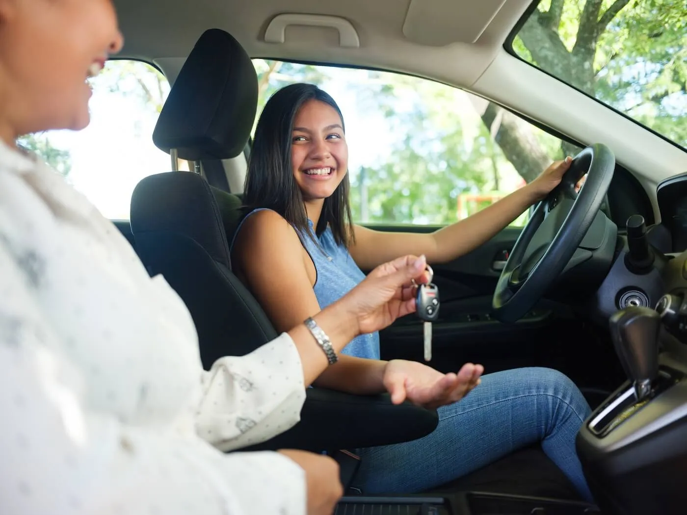 Smiling teenage girl sitting on a driver's seat and taking the car keys from the adult passenger