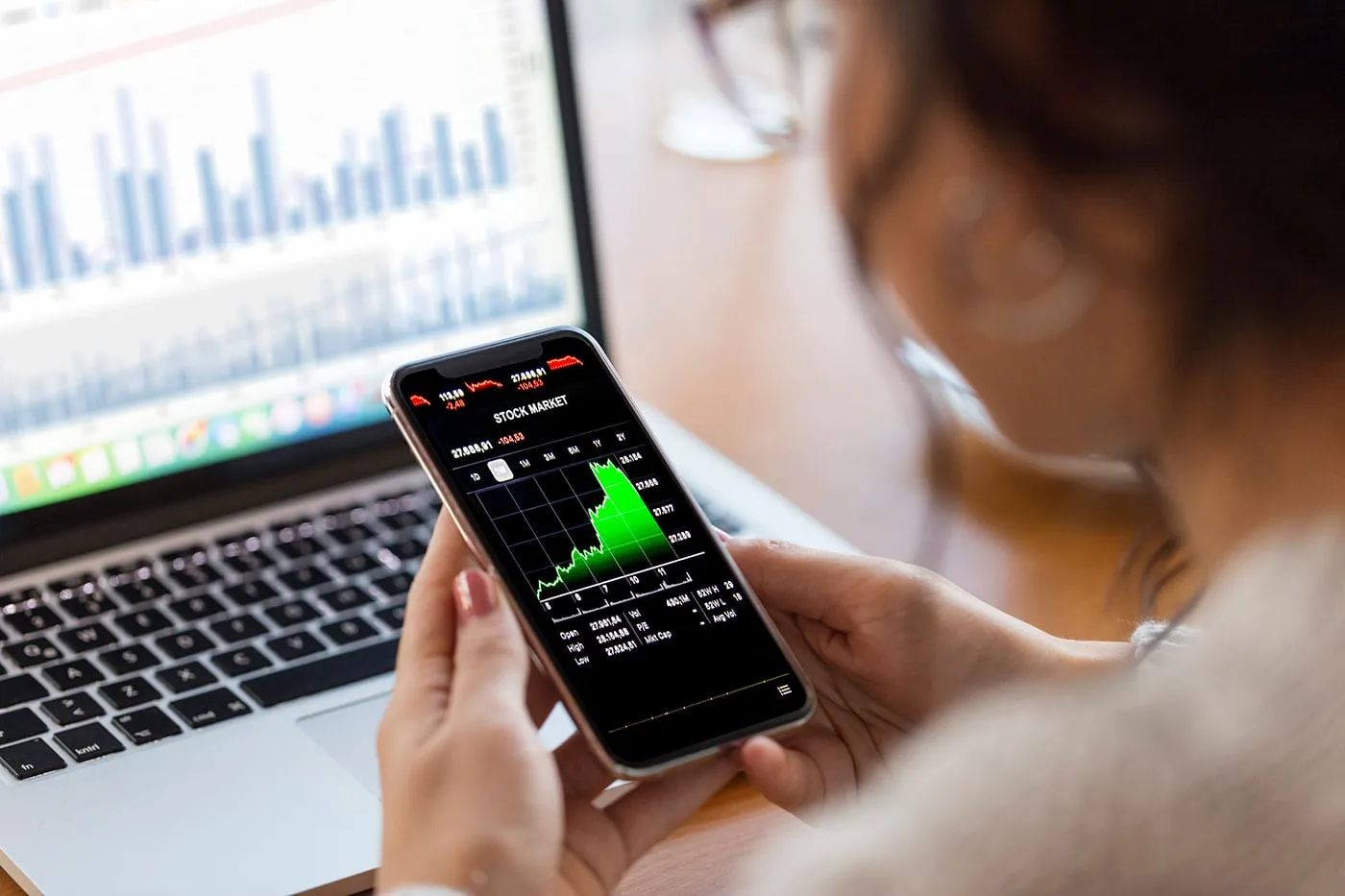 A woman looks at her phone showing a stock graph while her computer is in the background.