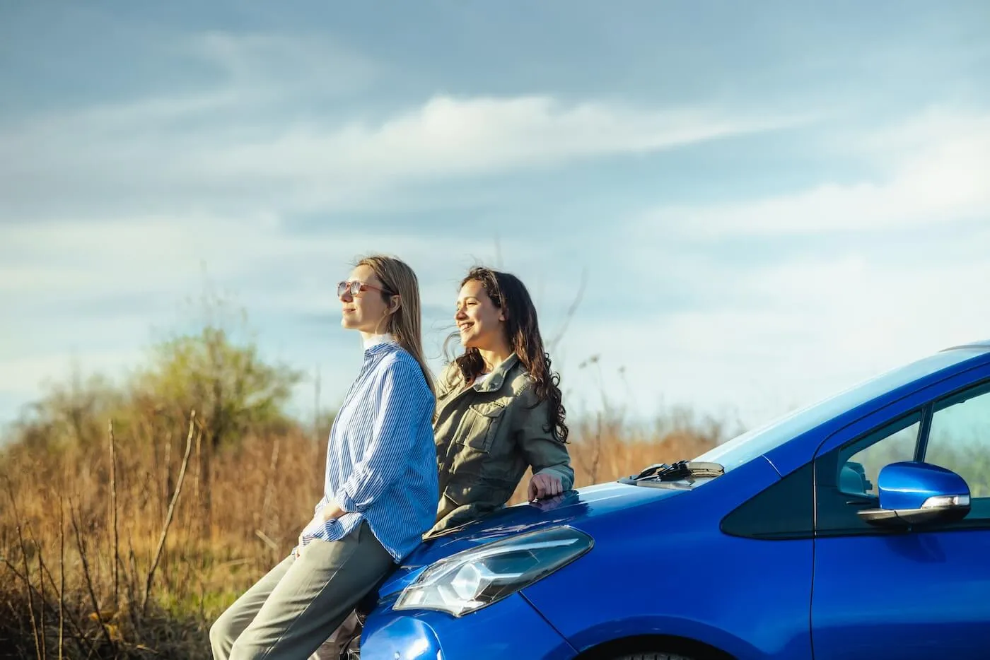 To smiling young women sitting on a blue car hood and observing the landscape