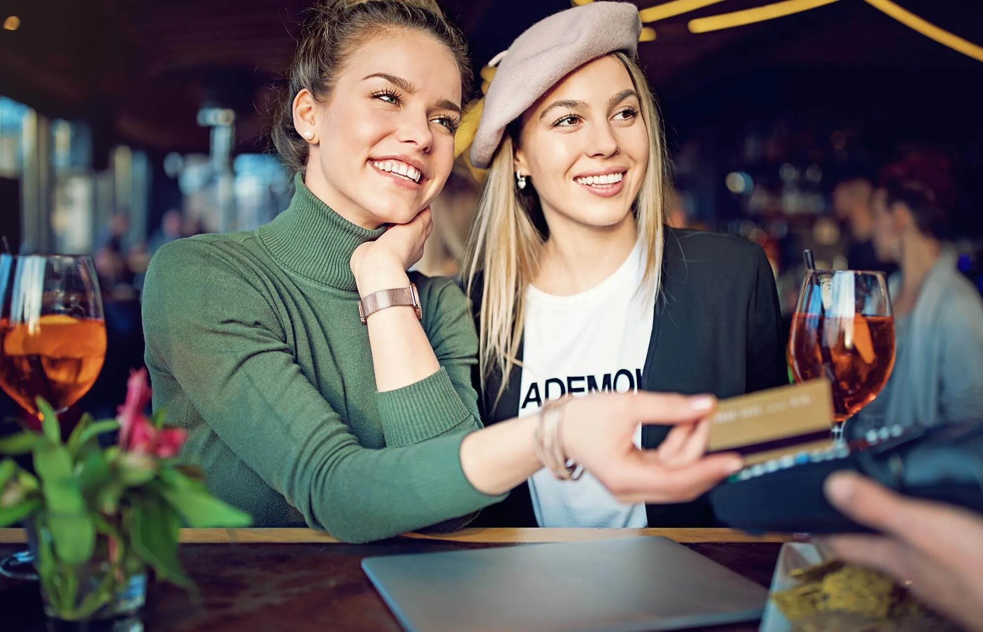 A woman wearing a green shirt smiles next to her friend while handing her credit card to the waiter to pay.