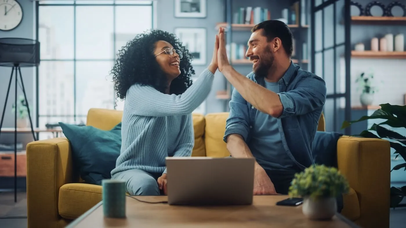 Happy couple giving high five while sitting on a couch in a living room with a laptop on a coffee table