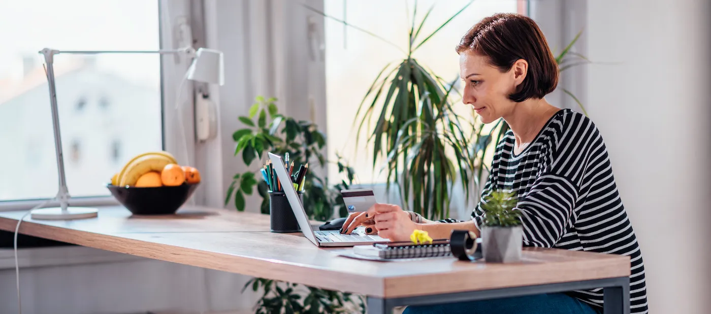 Woman using laptop to check credit score while holding credit card at the desk by the window