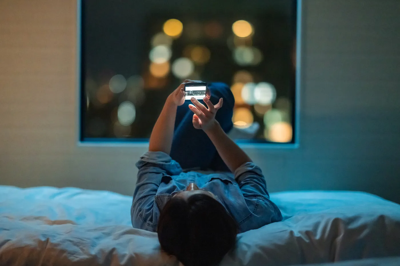 A woman is lying down on a bed and using a smart phone at night.