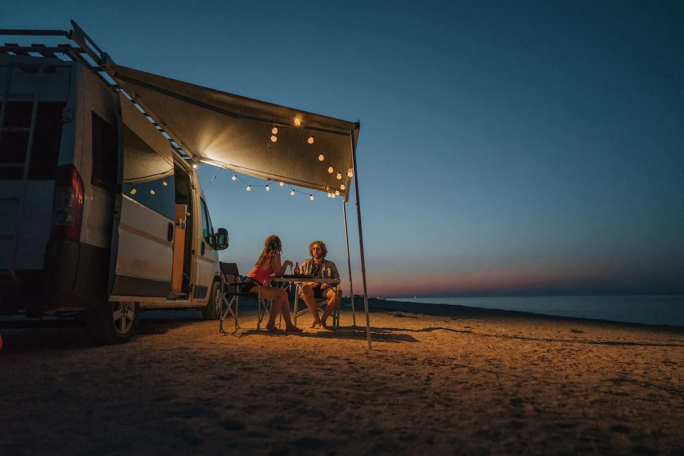 Young couple sitting outside their RV parked on the beach at sunset.