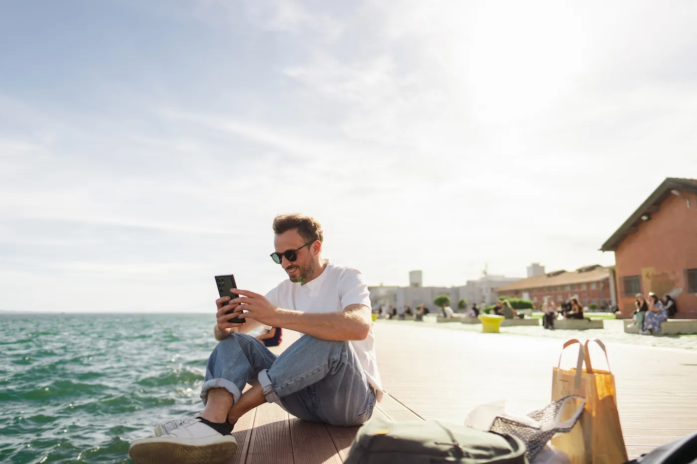 A man taking a break during the day, sitting on a pier and using his phone.