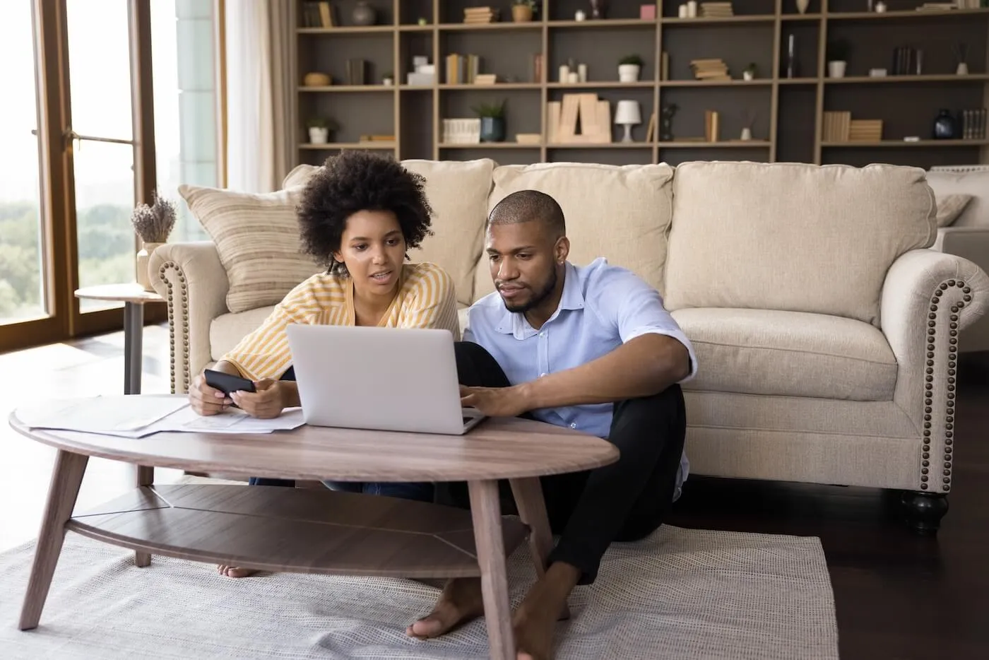 A couple is sitting on the floor in the living room while reviewing the bills and working on a laptop