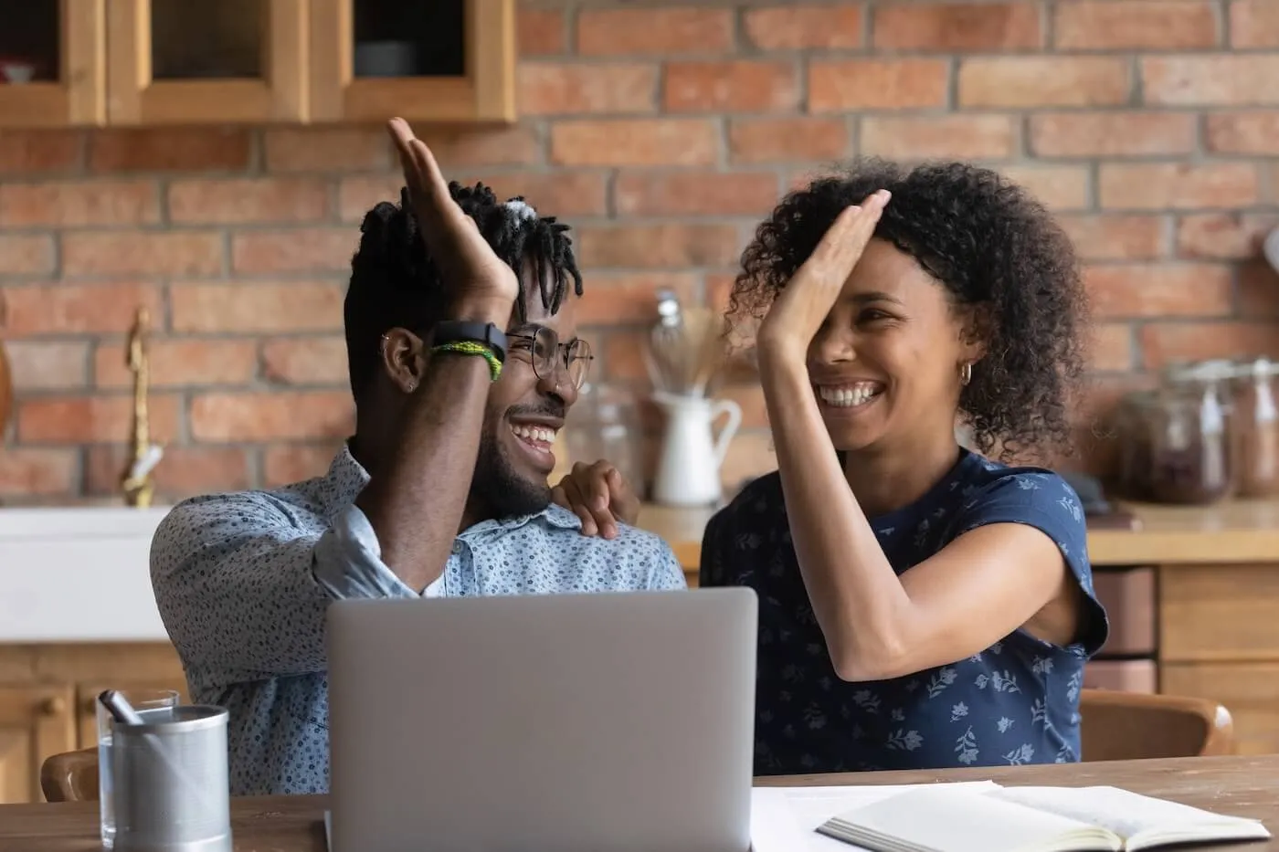 Happy couple giving high five to each other while sitting at the table with an open laptop and printouts