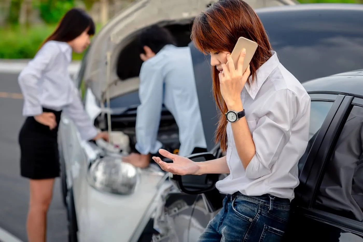 Woman calling the insurance company after a car collision, while couple on the background is looking under the hood of their car