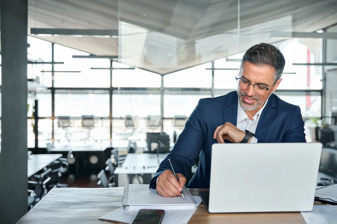 A smiling professional executive wearing suit sitting at desk in modern office working on laptop computer and writing notes.