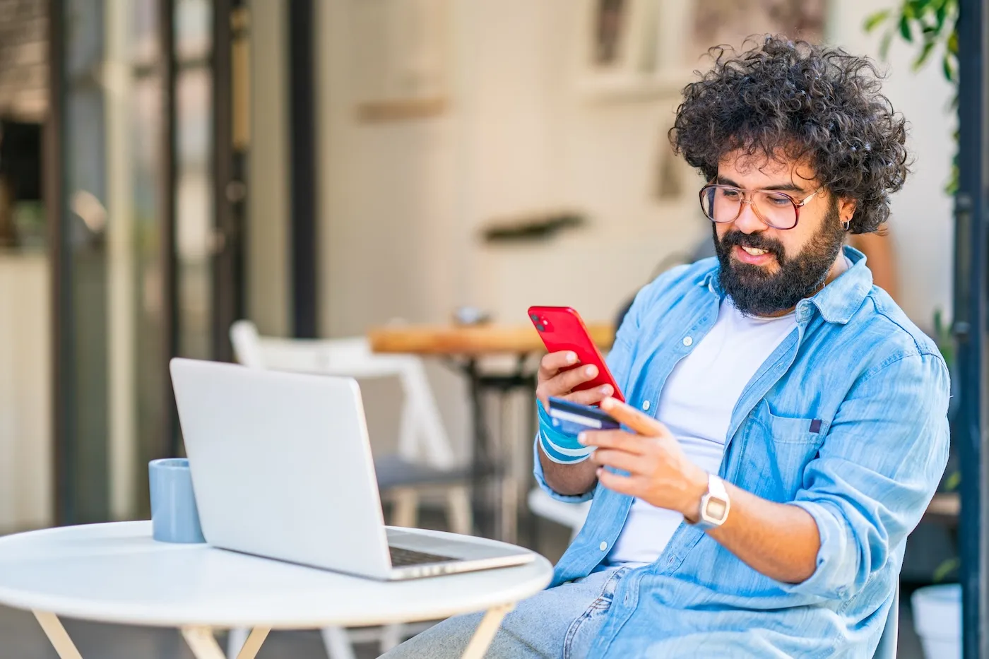 Curly haired man using mobile phone and credit card while sitting at an outdoor table.