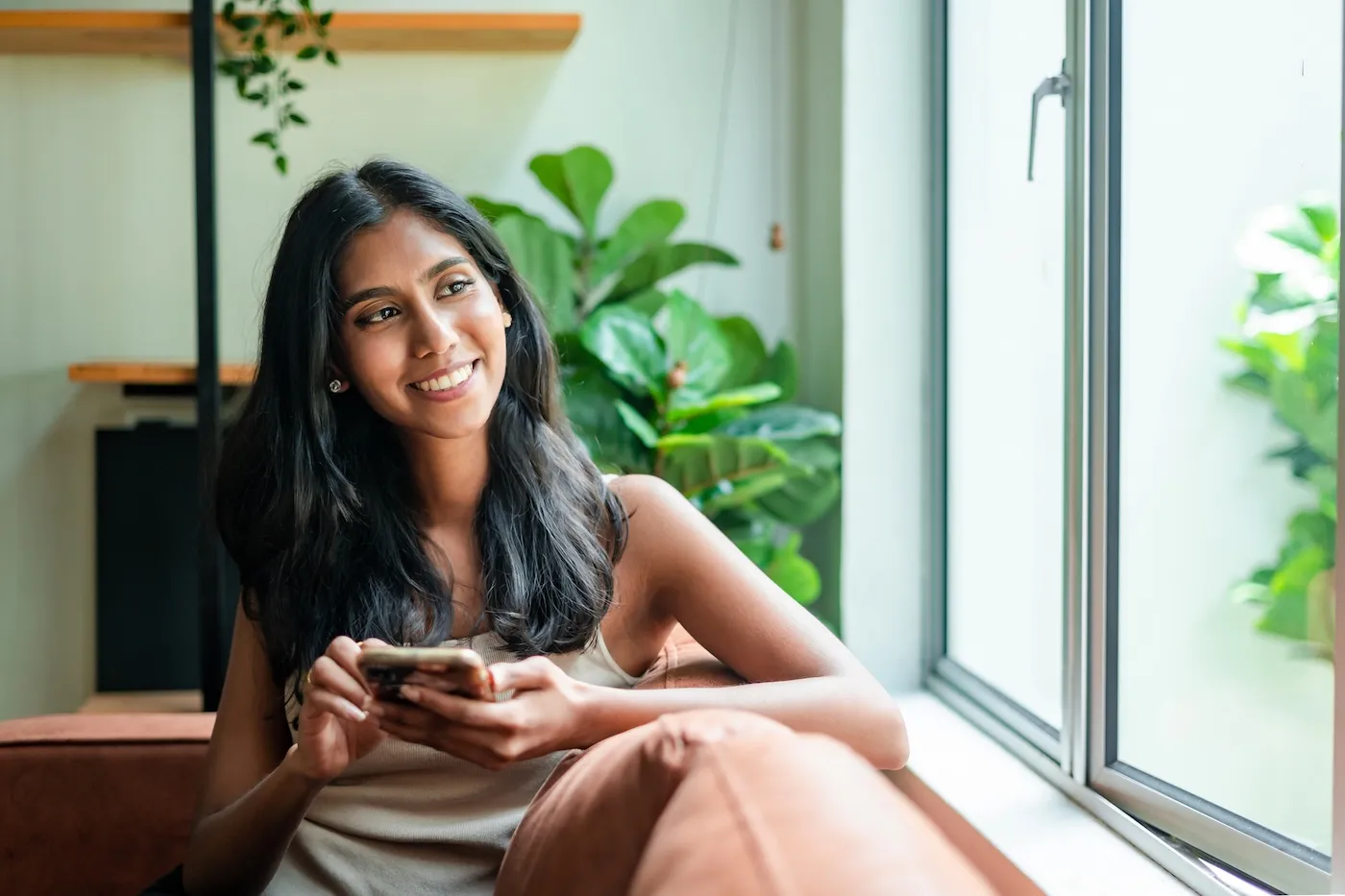 A woman relaxing on sofa at home, using smartphone and looking away with smile.